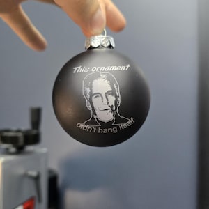 Puede incluir: Un adorno navideño negro con una imagen blanca de un hombre y el texto "This ornament didn't hang itself."