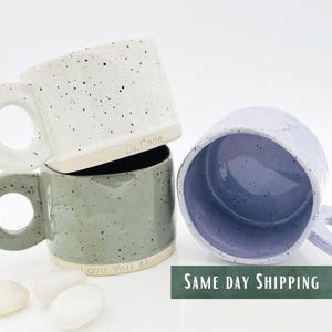 May include: Three ceramic mugs with speckled glaze. The white mug has the text "Dr. Cade" on the side. The green mug has the text "Love You Mom" on the side. The purple mug is empty.