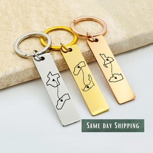 Könnte beinhalten: Drei Schlüsselanhänger mit rechteckigen Anhängern in Silber, Gold und Roségold. Jeder Schlüsselanhänger zeigt eine Kartenumriss mit einer gepunkteten Linie und Herzsymbolen, die eine Verbindung symbolisieren. Der Text "SAME DAY SHIPPING" ist sichtbar.