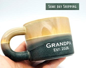Könnte beinhalten: Eine Keramik-Tasse mit gebogenem Henkel, die einen Farbverlauf von Beige bis Dunkelgrün aufweist. Auf der Tasse steht das Wort "GRANDPA" und das Jahr "EST. 2026". Die Tasse wird in einer Hand gehalten.