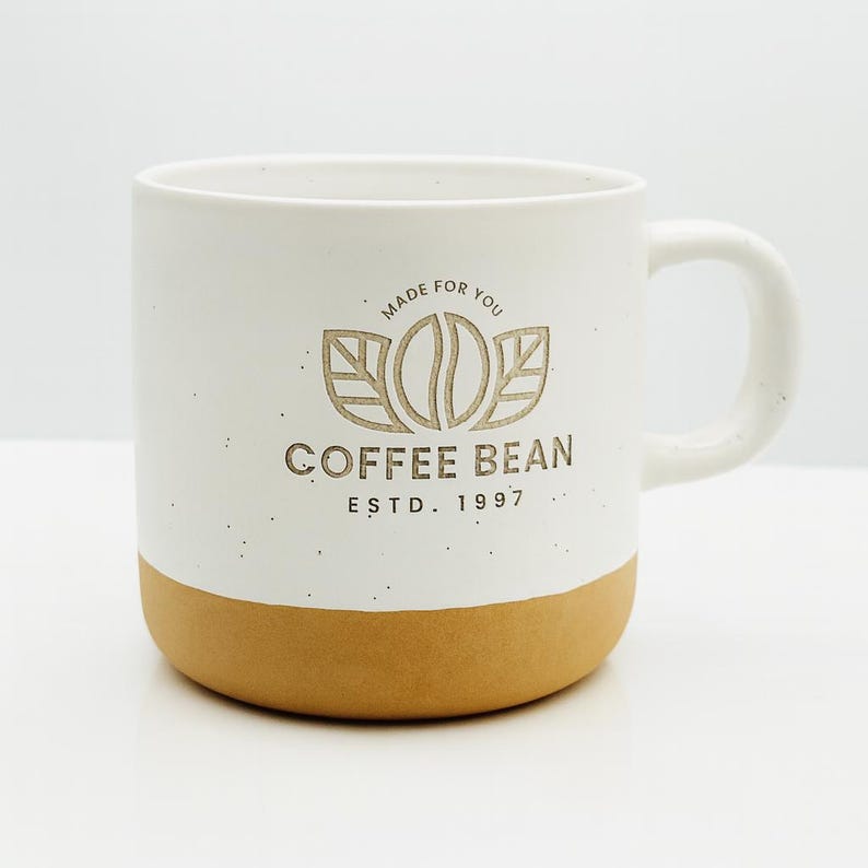 May include: A white ceramic mug with a speckled finish and a tan base. The front features a brown logo with a coffee bean icon, leaves, and the text MADE FOR YOU, COFFEE BEAN, and ESTD. 1997. The mug has a curved handle on the right side.