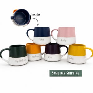 May include: A collection of six ceramic mugs with two-tone designs, featuring a coloured upper section and a white base. Each mug displays a unique word or phrase including "A morning," "What," "Smile," "Hey Handsome," "Hey There," and "Relax."