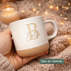 May include: A white ceramic mug with a speckled finish and a tan base. The mug features a large letter B in a serif font with the name Bella written in a cursive script across it. The mug has a curved handle on the right side.