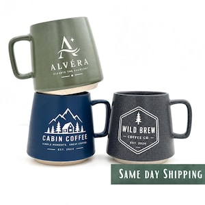 Könnte beinhalten: Drei Keramikbecher mit Henkeln. Der obere Becher ist salbeigrün mit dem weißen Text ALVÉRA ELEVATE THE EVERYDAY. Der untere linke Becher ist marineblau mit einer weißen Berg-Hütten-Grafik und CABIN COFFEE SIMPLE MOMENTS GREAT COFFEE EST. 2024. Der untere rechte Becher ist dunkelgrau mit einem weißen sechseckigen Logo und WILD BREW COFFEE CO. EST. 2024.