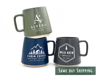 May include: Three ceramic mugs with handles. The top mug is sage green with the white text ALVÉRA ELEVATE THE EVERYDAY. The bottom left mug is navy blue with a white mountain cabin graphic and CABIN COFFEE SIMPLE MOMENTS GREAT COFFEE EST. 2024. The bottom right mug is dark gray with a white hexagon logo and WILD BREW COFFEE CO. EST. 2024.