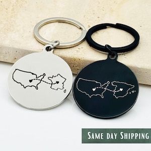 May include: Two circular keyrings, one silver and one black, each displaying a map outline of the United States and another country, linked by a dotted line with a heart. The silver keyring shows the US and France, the black one the US and Spain.