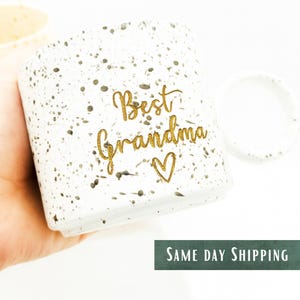 Pode incluir: Caneca de cerâmica branca com um padrão salpicado e as palavras "Best Grandma" em dourado, com um coração dourado por baixo. A caneca tem formato arredondado e uma alça. O texto "Same Day Shipping" está na parte inferior.