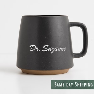 May include: A black ceramic mug with a wide base and a curved handle. The mug features the text "Dr. Suzanne" in a white script font. The base of the mug is a light brown colour.