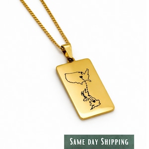 May include: Gold-colored rectangular pendant necklace with a chain. The pendant features a black outline of the United States and Great Britain, connected by a line with hearts. The necklace is on a white background.