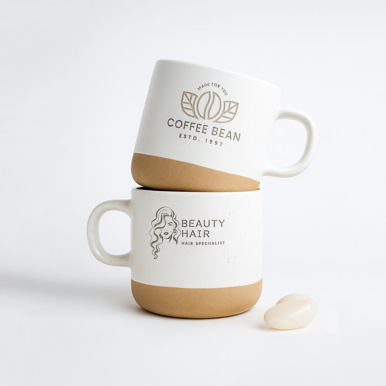 May include: Two stacked ceramic mugs with a white glazed upper section and a natural tan base. The top mug features a coffee bean logo with the text COFFEE BEAN ESTD. 1997. The bottom mug displays a line art illustration of a person with wavy hair.