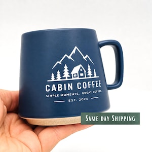 Może przedstawiać: Matowy, granatowy kubek ceramiczny z białą grafiką przedstawiającą chatę pośród sosen i gór. Napis głosi CABIN COFFEE, SIMPLE MOMENTS. GREAT COFFEE. EST. 2024. Kubek posiada solidne ucho oraz podstawę w naturalnym, beżowym odcieniu.