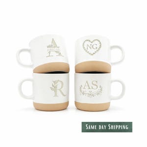 May include: Four white ceramic mugs stacked in two pairs. Each mug has a tan base and a white upper section with a handle. The mugs are personalized with initials and names in a light tan color.