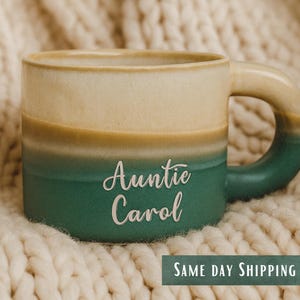 Puede incluir: Una taza de cerámica de dos tonos con la parte superior crema, una banda amarillo-marrón y una base verde-turquesa. La taza presenta el texto "Auntie Carol" en cursiva. El asa y la base son verde-turquesa. La taza está sobre una superficie de punto crema.