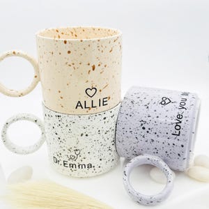 Pode incluir: Três canecas de cerâmica salpicadas com grandes alças circulares. Uma é bege com salpicos castanhos e o nome "Allie" e um coração. Outra é branca com salpicos pretos e o nome "Dr. Emma" e um estetoscópio. A terceira é cinzenta com as palavras "Love you Mom".