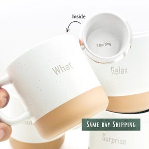 May include: White ceramic mugs with a speckled finish and a tan base. The mugs feature the text What, Relax, and Surprise on the exterior. One mug shows the interior with the text A morning printed at the bottom. The mugs have a handle.