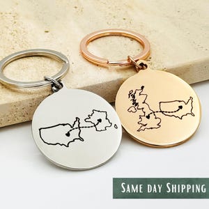 Puede incluir: Dos llaveros con colgantes redondos, uno plateado y otro dorado. Cada colgante presenta un contorno de mapa de Estados Unidos y Reino Unido, conectados por una línea punteada. El texto "SAME DAY SHIPPING" está en la parte inferior.