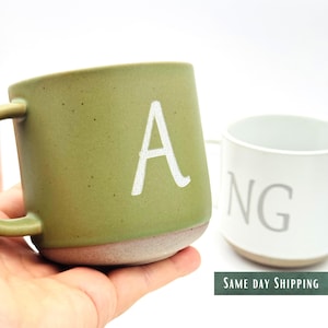 Puede incluir: Dos tazas de cerámica, una verde oliva y otra blanca. La taza verde tiene una letra blanca "A". La taza blanca tiene las letras "NG". Ambas tazas tienen un asa. Las tazas están sobre un fondo blanco.