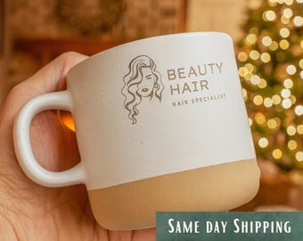 May include: A white ceramic mug with a speckled finish and a tan base. The front features a dark brown line art illustration of a person with wavy hair, accompanied by the text BEAUTY HAIR and HAIR SPECIALIST in a clean, sans-serif font.