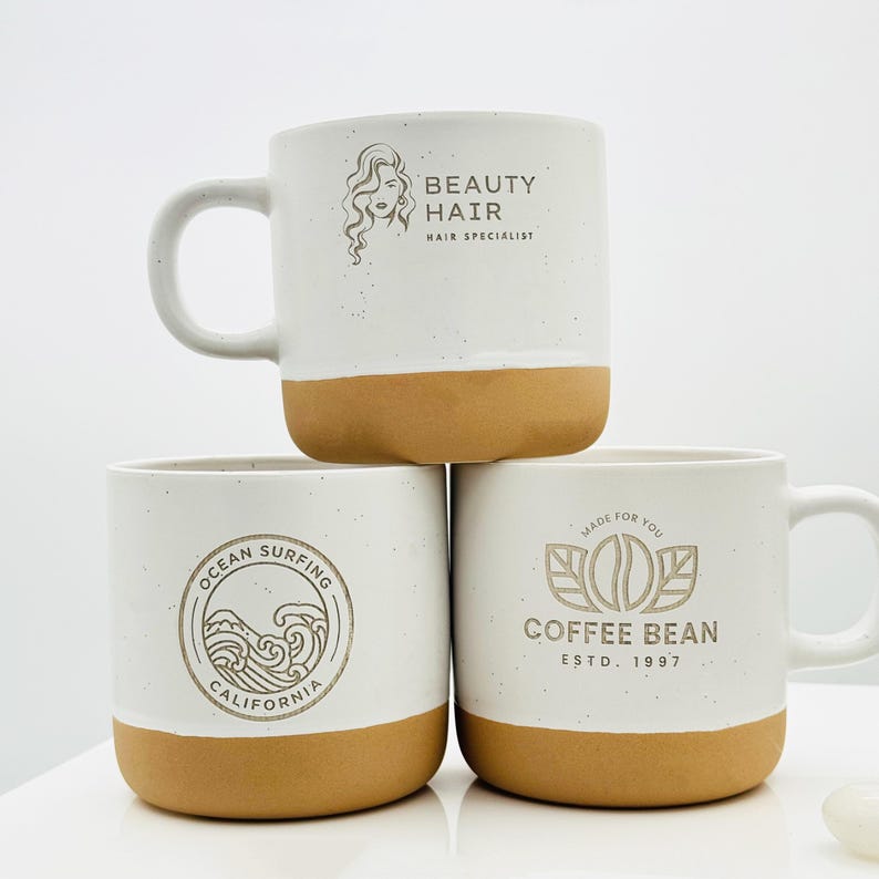 May include: Three ceramic mugs with a speckled white upper section and a solid tan base. The top mug features a Beauty Hair logo. The bottom left mug displays an Ocean Surfing California graphic. The bottom right mug shows a Coffee Bean logo.