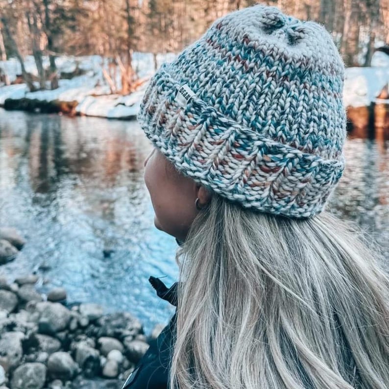 May include: A woman wearing a multi-coloured knitted beanie with a white label that says "BRIAR". The beanie is made of a soft, chunky yarn and has a slouchy fit. The woman is standing in front of a river with rocks and snow in the background.