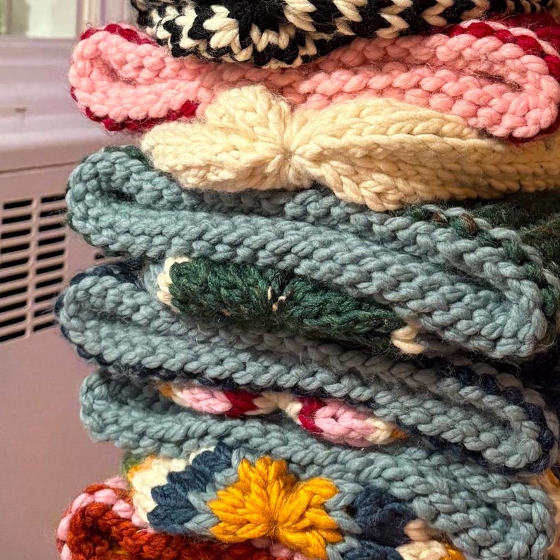 May include: A stack of colourful knitted hats with different patterns and textures. The hats are made of yarn in shades of pink, white, blue, green, and yellow.
