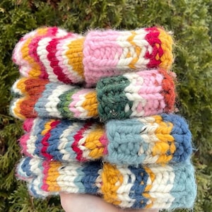 May include: A stack of four thick, hand-knitted wool mittens featuring vibrant, multi-coloured chevron patterns. Each mitten displays a unique combination of pink, yellow, red, green, blue, and cream yarn, showcasing a chunky, textured knit stitch.