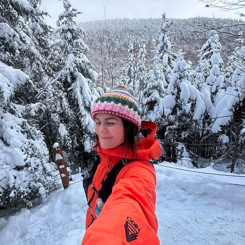 May include: A person wearing a bright orange jacket with a black logo that says "ODIN" is standing in a snowy forest. They are wearing a pink and green striped knitted hat.