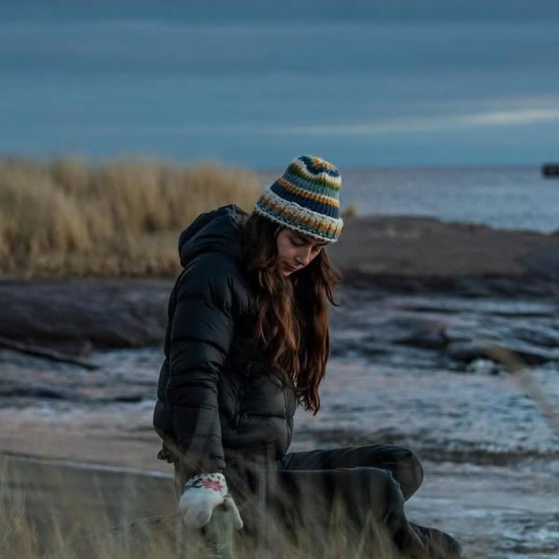 May include: A woman wearing a black puffer jacket with a hood and a colourful striped knit beanie. She is sitting on a rocky shore with a body of water in the background.