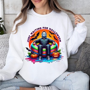 May include: White sweatshirt with a graphic of a masked figure sitting in a colorful inflatable ring. The text "Just Waiting for Halloween" is written in a circle around the figure.
