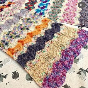 Pode incluir: Uma manta de crochê multicolorida com um padrão ondulado. A manta é feita com diferentes tons de fio rosa, roxo, azul, laranja e branco.