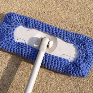 Puede incluir: Una cabeza de fregona de crochet azul unida a un mango de fregona blanco. La cabeza de fregona está hecha de un hilo suave y texturizado y tiene un borde decorativo.