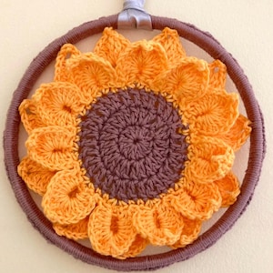 May include: A crocheted sunflower wall hanging with a brown center and yellow petals. The sunflower is attached to a brown wooden hoop.