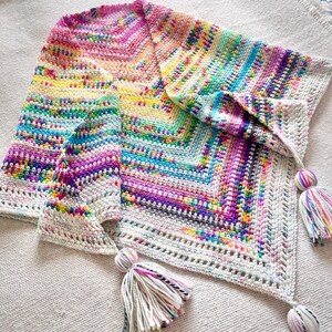 Pode incluir: Um xale de crochê colorido com um padrão de arco-íris e franjas brancas. O xale é feito com uma variedade de cores, incluindo rosa, roxo, azul, verde, amarelo e laranja. As franjas são feitas de fio branco e são presas à borda inferior do xale.