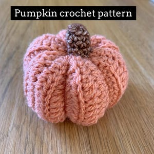 Puede incluir: Una calabaza de ganchillo de color melocotón con un tallo de ganchillo marrón.  "Pumpkin crochet pattern" está escrito encima de la calabaza.