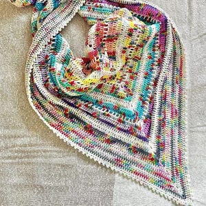 Pode incluir: Um xale de crochê colorido com uma borda branca e uma borda festonada. O xale é feito com uma variedade de cores, incluindo rosa, azul, verde, amarelo e roxo.