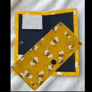 May include: A handmade wallet with a yellow bee pattern on the front flap. The wallet is open, revealing a dark interior with a white, scalloped-edge patch. The edges are trimmed with yellow fabric. A black snap closure is visible.