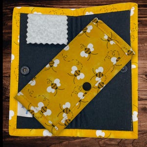 May include: A yellow and black glasses case with a bee pattern, resting on a dark gray background. The case is inside a larger yellow and white quilted holder with a snap closure. A white cleaning cloth is visible.
