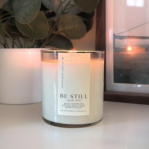May include: A lit, clear glass candle with a white label that reads "BE STILL" and "Psalm 46:10". The candle is 9 oz (255 g) and sits in front of a framed picture and a white pot with a plant.