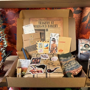 May include: A cardboard box filled with mystery-themed items including a newspaper titled Tragedy at Marigold Bakery, a reading list card, a brown envelope, a bookmark, a Tazo tea bag, a pair of patterned socks, stickers, and a wooden pipe.