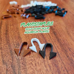 Three plant anchors, one brown, one white, and one black, stand upright on a wooden surface in the foreground, with many more blurred in the background, and green text that reads "PLANTSTAPLES HOLD DOWN YOUR AQUARIUM PLANTS!"