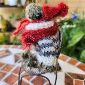 May include: A small knitted toy animal wearing a red scarf and hat, sitting on a metal chair. The toy is made of brown, white, and blue yarn. The chair is on a tiled floor.