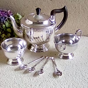 May include: A silver tea set comprising a teapot, sugar bowl, creamer jug, and small spoons. The teapot features a black handle and lid knob. The set is arranged on a light-coloured surface.