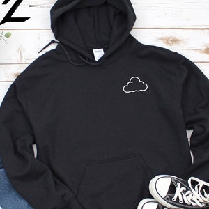 Peut inclure: Sweat-shirt à capuche noir avec un graphique de nuage blanc sur le devant. Le sweat-shirt est présenté sur un fond en bois blanc avec un jean et des baskets.