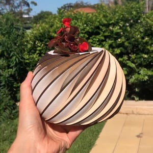Op de afbeelding: Een houten plantenbak met een swirl-ontwerp, geschilderd in wit en bruin. De plantenbak bevat een kleine plant met rode bloemen.