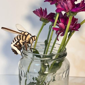 Può includere: Una statuetta di ape in legno con strisce bianche e nere si trova sul bordo di un vaso di vetro trasparente. Il vaso è pieno di fiori viola con steli verdi.