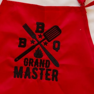 May include: Red BBQ apron with black embroidered text that reads "BBQ Grand Master" with a crossed spatula and fork design.