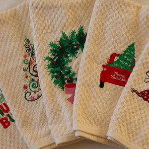 Op de afbeelding: Een verzameling crèmekleurige keukendoeken met wafelweefsel en geborduurde kerstontwerpen. Ontwerpen omvatten een kerstboom, een rode vrachtwagen met een boom, een sneeuwpop en feestelijke tekst. Elke handdoek heeft een uniek ontwerp.