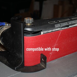 Pu&ograve; includere: Una levigatrice a nastro rossa e nera con una porta di aspirazione della polvere e una base in plastica nera. La levigatrice ha una manopola nera sul lato e il testo "compatible with stop" &egrave; scritto sulla levigatrice.