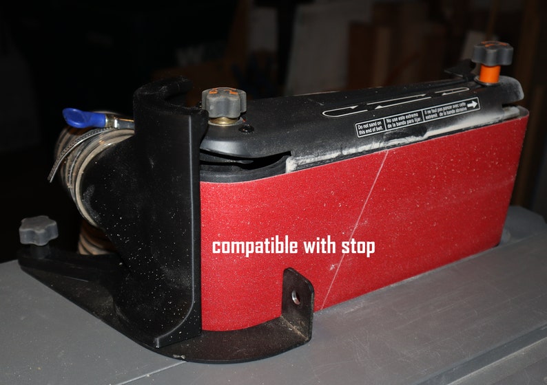 Pu&ograve; includere: Una levigatrice a nastro rossa e nera con una porta di aspirazione della polvere e una base in plastica nera. La levigatrice ha una manopola nera sul lato e il testo "compatible with stop" &egrave; scritto sulla levigatrice.