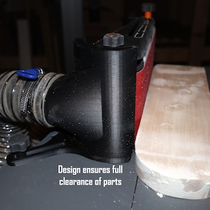 Pu&ograve; includere: Un sistema di raccolta della polvere nero con un tubo collegato a una levigatrice. La levigatrice ha una cinghia abrasiva rossa ed &egrave; fissata a una base nera. Il testo "Design ensures full clearance of parts" &egrave; visibile sotto la levigatrice.
